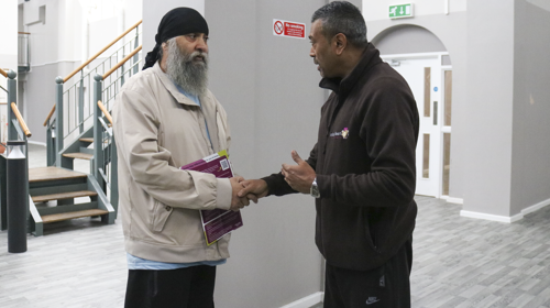 Two Individuals Shaking Hands Inside A Building With Stairs And A Railing In The Background