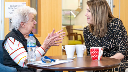 Two Women Speaking While Sat At A Table With Mugs