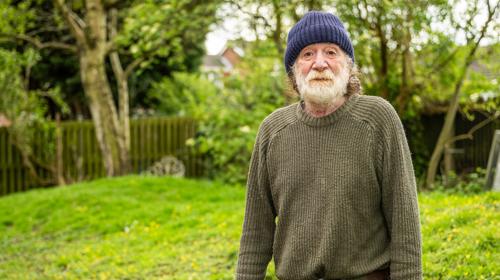 Man With A Beard Standing Against A Garden Backdrop Wearing A Hat And Green Jumper