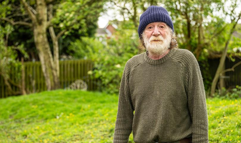 Man With A Beard Standing Against A Garden Backdrop Wearing A Hat And Green Jumper