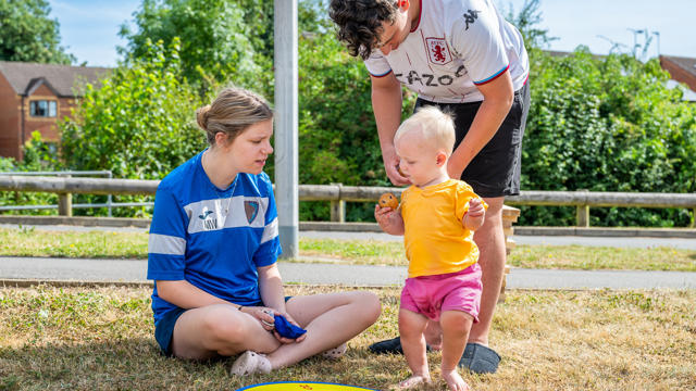 Two Individuals In Sports Jerseys Watch A Toddler Taking Steps On A Colorful Play Mat Outdoors