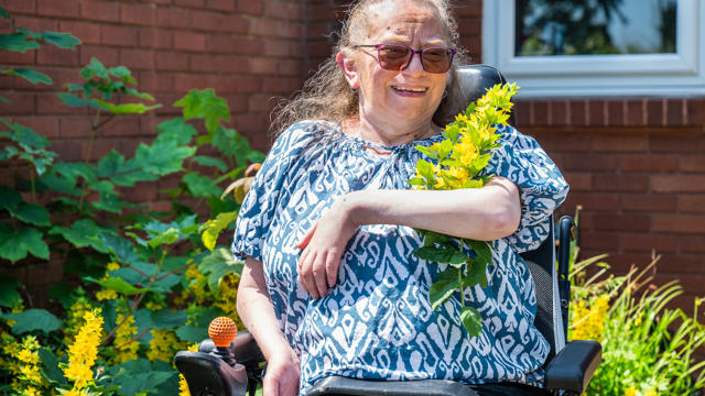 An Individual In A Wheelchair Holding A Bunch Of Yellow Flowers And Plants And Smiling, Outdoors On A Sunny Day