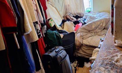 Cluttered Bedroom With Clothes On A Rail And Piled On The Bed, A Suitcase And Bags On The Floor, And Toiletries On A Nearby Surface