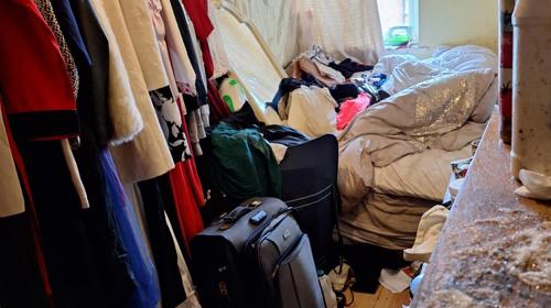 Cluttered Bedroom With Clothes On A Rail And Piled On The Bed, A Suitcase And Bags On The Floor, And Toiletries On A Nearby Surface