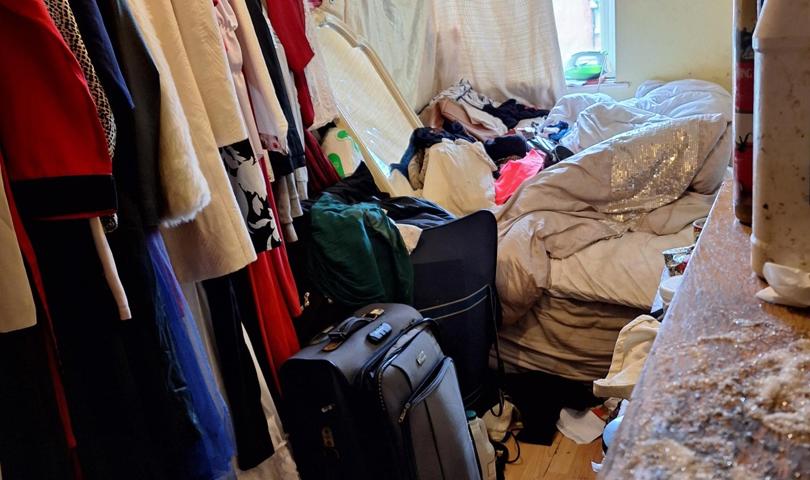 Cluttered Bedroom With Clothes On A Rail And Piled On The Bed, A Suitcase And Bags On The Floor, And Toiletries On A Nearby Surface
