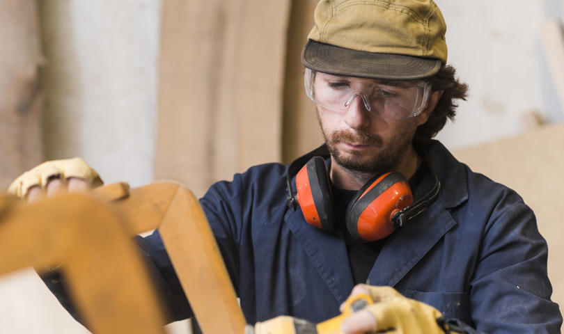 A Person In A Workshop Wearing Safety Gear, Including Goggles And Ear Protection, Is Attentively Using A Power Tool On A Piece Of Wood