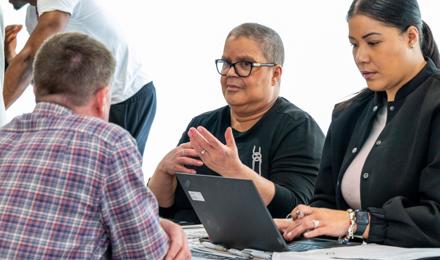 Three People Engaged In A Serious Discussion Around A Table, With Laptops And Documents In Front Of Them. Two Individuals Are Visible Facing The Camera