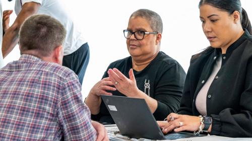Three People Engaged In A Serious Discussion Around A Table, With Laptops And Documents In Front Of Them. Two Individuals Are Visible Facing The Camera