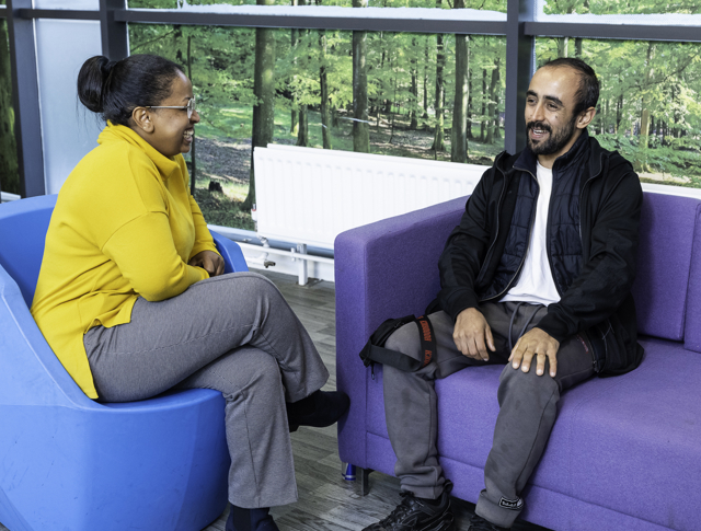 Two People Engaged In A Conversation While Sitting On Colorful Chairs In A Room With Large Windows Overlooking A Forest