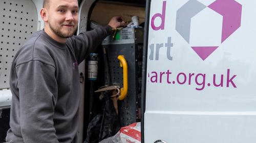 A Person Smiling, Standing Next To A White Van Marked With A Midland Heart Logo. The Van's Door Is Open Showing Tools And Equipment Inside