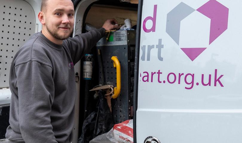A Person Smiling, Standing Next To A White Van Marked With A Midland Heart Logo. The Van's Door Is Open Showing Tools And Equipment Inside