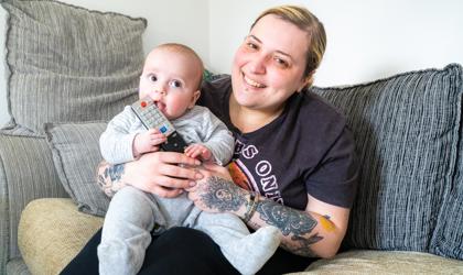 Baby And Mother Sitting On Sofa Smiling At The Camera While Baby Chews On Remote Control
