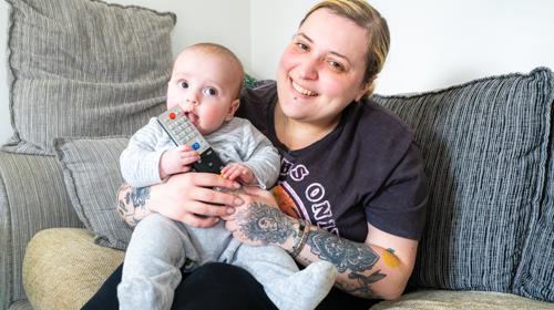 Baby And Mother Sitting On Sofa Smiling At The Camera While Baby Chews On Remote Control