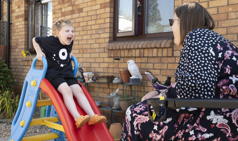 A Child Slides Down A Plastic Slide, Excitedly Interacting With An Adult Seated Nearby, In A Sunny Backyard Setting With A Small Bird Perched Close By
