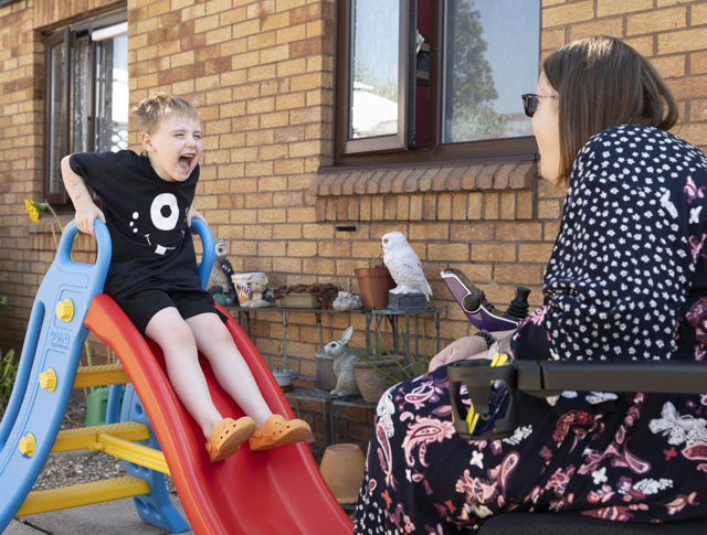 A Child Slides Down A Plastic Slide, Excitedly Interacting With An Adult Seated Nearby, In A Sunny Backyard Setting With A Small Bird Perched Close By
