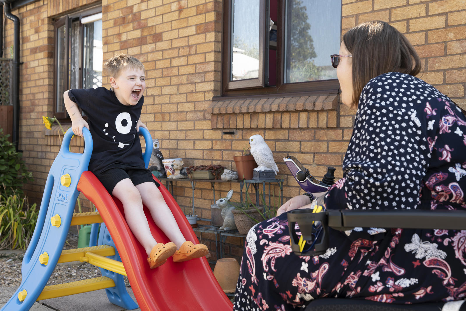 A Child Slides Down A Plastic Slide, Excitedly Interacting With An Adult Seated Nearby, In A Sunny Backyard Setting With A Small Bird Perched Close By