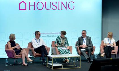 A Stage Stage Photo With A Panel Of Five People Seated And Speaking With A Big Screen In The Background Which Says HOUSING