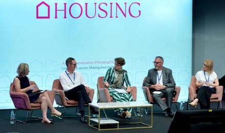 A Stage Stage Photo With A Panel Of Five People Seated And Speaking With A Big Screen In The Background Which Says HOUSING