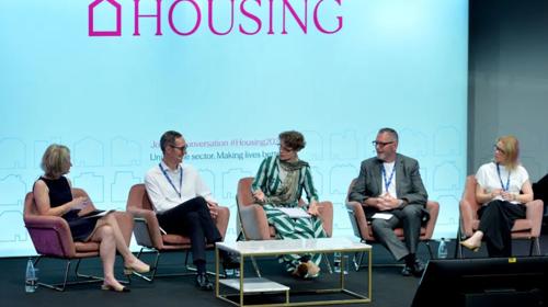 A Stage Stage Photo With A Panel Of Five People Seated And Speaking With A Big Screen In The Background Which Says HOUSING