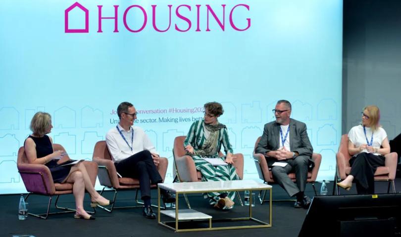 A Stage Stage Photo With A Panel Of Five People Seated And Speaking With A Big Screen In The Background Which Says HOUSING