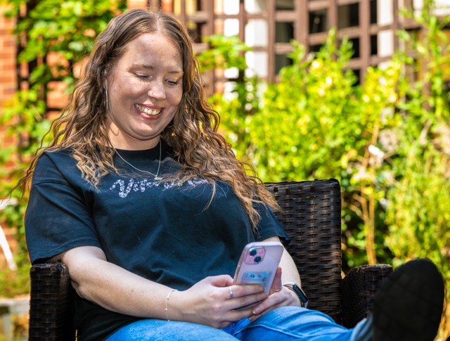 Person Sitting On A Wicker Chair Outdoors, Smiling While Looking At A Smartphone. They Are Wearing A Casual Blue Shirt And Jeans, Surrounded By A Garden With Brick Buildings In The Background