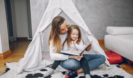 Mother With Little Daughter At Home Sitting Reading A Book Under A Homemade Tent