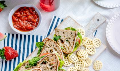 Sandwiches On A Cutting Board With Crisps Scattered Beside And A Red Dip In A Picnic Setting