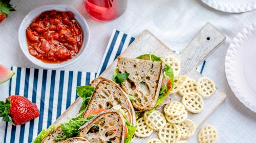 Sandwiches On A Cutting Board With Crisps Scattered Beside And A Red Dip In A Picnic Setting