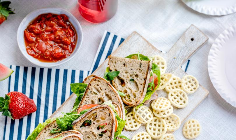 Sandwiches On A Cutting Board With Crisps Scattered Beside And A Red Dip In A Picnic Setting