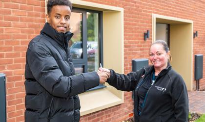 Two People Smiling At Camera While One Is Giving The Other Something Stood Outside A New House