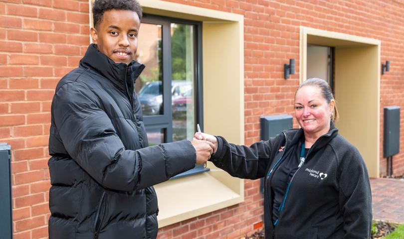 Two People Smiling At Camera While One Is Giving The Other Something Stood Outside A New House