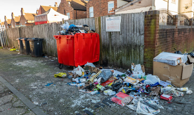 An Outdoor Area With Red And Black Trash Bins In Front Of A Wooden Fence. Trash, Including Cardboard, Plastic, And Other Debris, Is Scattered On The Ground