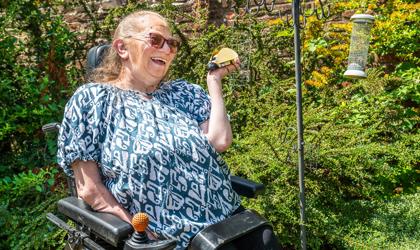 Lady Sitting In Wheelchair Out In A Garden Smiling At A Plastic Ladybird In Her Hand