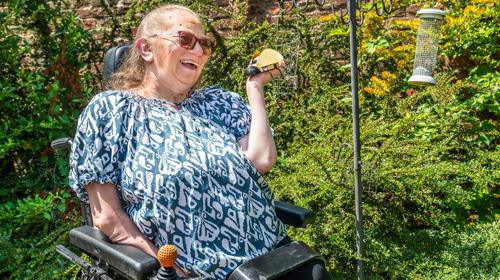 Lady Sitting In Wheelchair Out In A Garden Smiling At A Plastic Ladybird In Her Hand