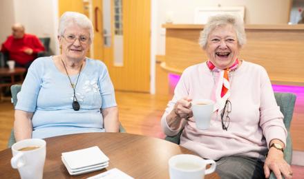 Two Elderly Individuals Smiling And Holding Mugs At A Table In A Bright Room, With Other People Visible In The Background