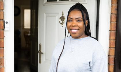 Woman With Long Hair In Braids Stood At Her Front Door Smiling At The Camera
