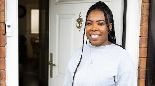 Woman With Long Hair In Braids Stood At Her Front Door Smiling At The Camera