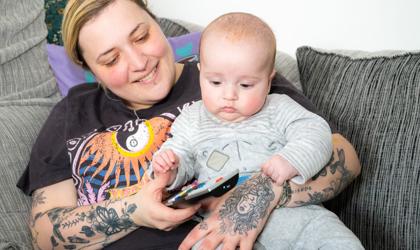 A Person With Tattooed Arms Is Sitting On A Sofa, Smiling At A Baby Who Is Holding A TV Remote. They Are Both Sitting Comfortably Among Cushions