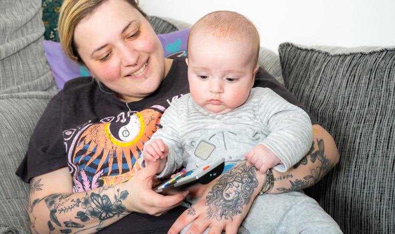 A Person With Tattooed Arms Is Sitting On A Sofa, Smiling At A Baby Who Is Holding A TV Remote. They Are Both Sitting Comfortably Among Cushions
