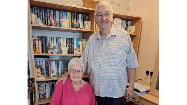 Older Couple Posing For Camera With Man Standing And Woman Sat On A Chair Against A Bookcase