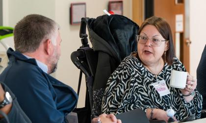 Person With A Name Tag Labelled 'Visitor' Holding A Coffee Mug And Engaging In Conversation With Another Individual At A Table During A Meeting