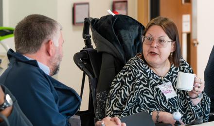 Person With A Name Tag Labelled 'Visitor' Holding A Coffee Mug And Engaging In Conversation With Another Individual At A Table During A Meeting