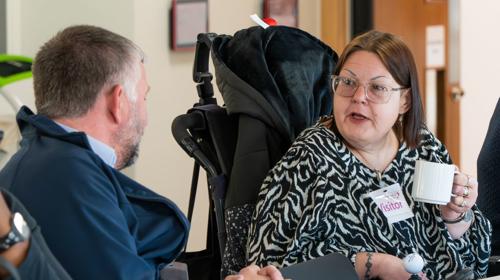 Person With A Name Tag Labelled 'Visitor' Holding A Coffee Mug And Engaging In Conversation With Another Individual At A Table During A Meeting