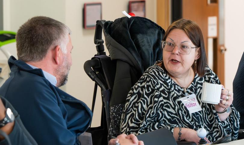 Person With A Name Tag Labelled 'Visitor' Holding A Coffee Mug And Engaging In Conversation With Another Individual At A Table During A Meeting