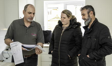 Three Individuals Examine A Document Together Inside A Kitchen, With One Person From Mybuilder Assisting The Other Two