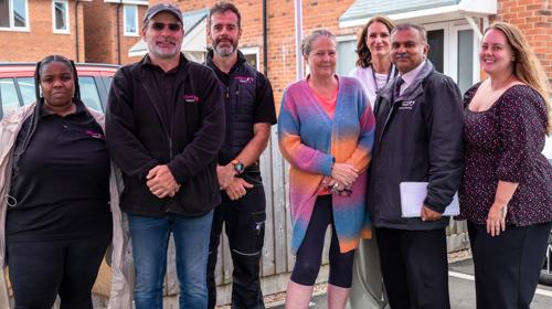 A Diverse Group Of Seven People Standing Together Under A Canopy, Smiling. They Are Dressed In Casual And Business Attire With Brick Houses In The Background