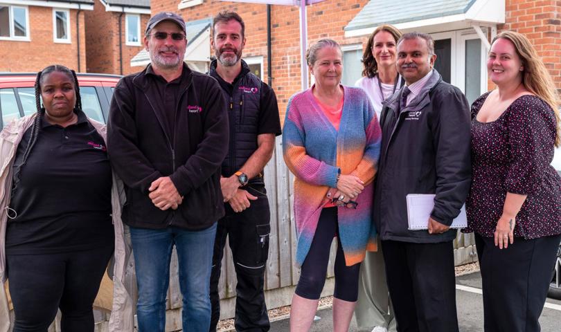 A Diverse Group Of Seven People Standing Together Under A Canopy, Smiling. They Are Dressed In Casual And Business Attire With Brick Houses In The Background