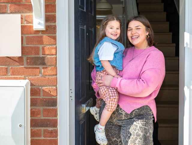 The Image Shows Two Smiling Individuals At The Entrance Of A Red Brick Building. One Individual Is Holding A Young Child, And They Both Appear Happy