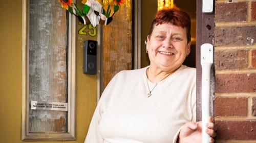 A Tenant Standing Outside Against Their Yellow Door, Holding A Handrail And Smiling