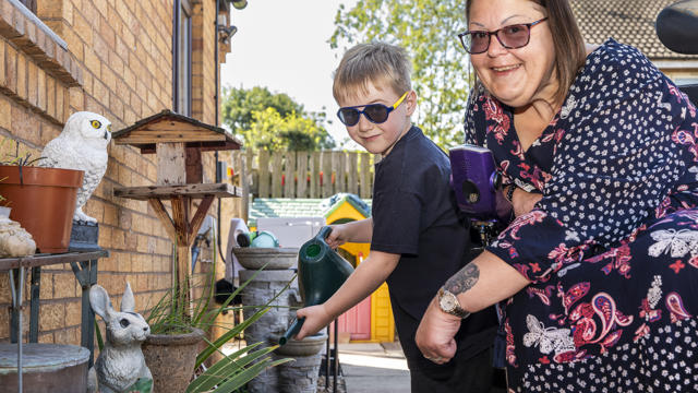 Two People Happily Gardening Together In A Sunny Backyard, Surrounded By Plants And Garden Decorations. The Child Is Watering Plants With A Watering Can, While The Adult, Wearing Sunglasses, Smiles At The Camera